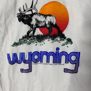 May include: White t-shirt featuring a black and white elk silhouette on a grassy hill, with an orange and red sun in the background. The word "Wyoming" is printed in blue, with additional text below.