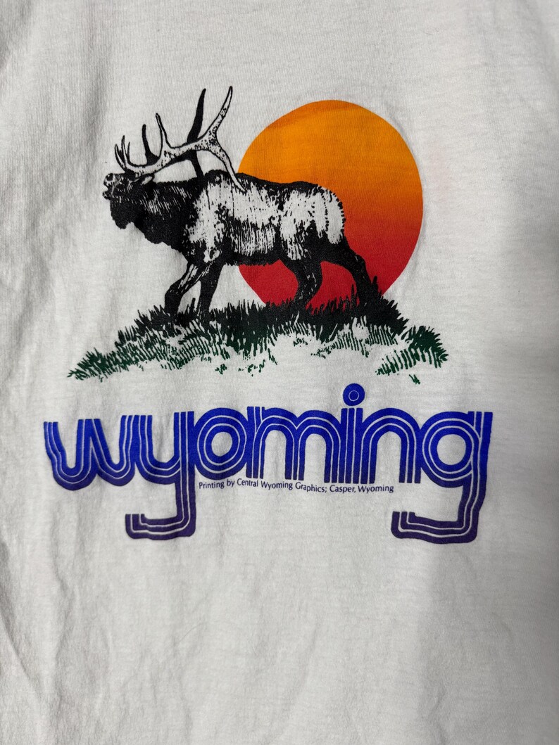 May include: White t-shirt featuring a black and white elk silhouette on a grassy hill, with an orange and red sun in the background. The word "Wyoming" is printed in blue, with additional text below.