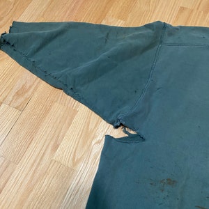May include: A dark green sweatshirt with a distressed look. The sweatshirt has several holes and tears in the fabric, and there are some brown stains on the fabric.