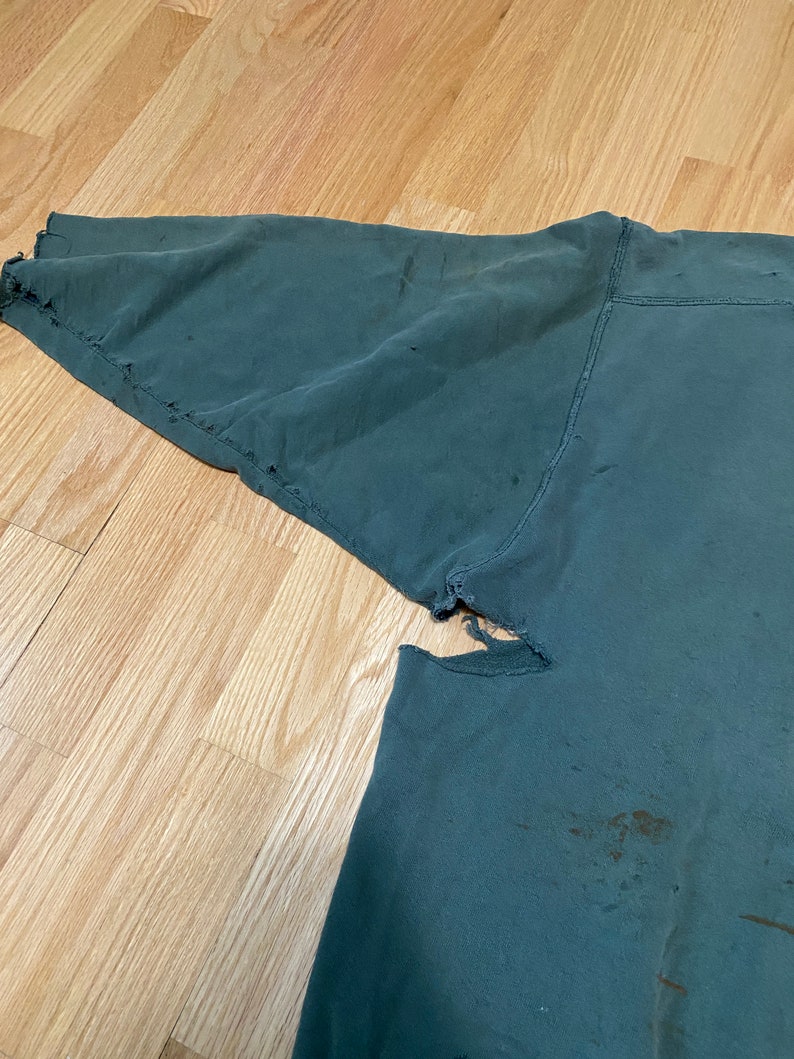 May include: A dark green sweatshirt with a distressed look. The sweatshirt has several holes and tears in the fabric, and there are some brown stains on the fabric.