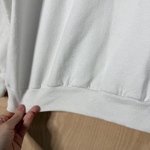 May include: Close-up of a white sweatshirt with a ribbed hem and cuffs. The fabric is slightly wrinkled, and the garment appears to be made of a soft material. The image shows a portion of the sweatshirt.