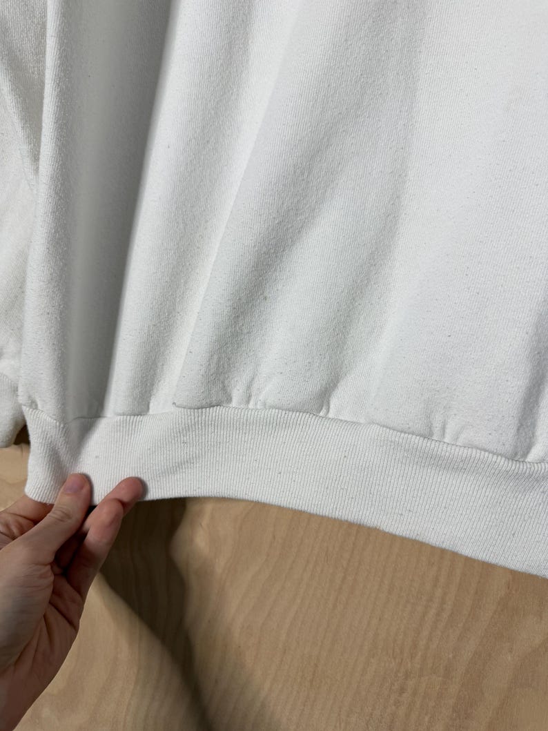 May include: Close-up of a white sweatshirt with a ribbed hem and cuffs. The fabric is slightly wrinkled, and the garment appears to be made of a soft material. The image shows a portion of the sweatshirt.