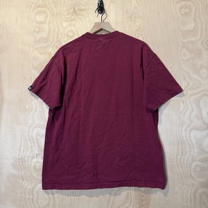 K&ouml;nnte beinhalten: Ein weinrotes T-Shirt mit kurzen &Auml;rmeln h&auml;ngt an einem Holzkleiderb&uuml;gel. Das Shirt besteht aus einem weichen Material und hat ein schlichtes, klassisches Design. Der Hintergrund ist eine hellfarbige Holzoberfl&auml;che.