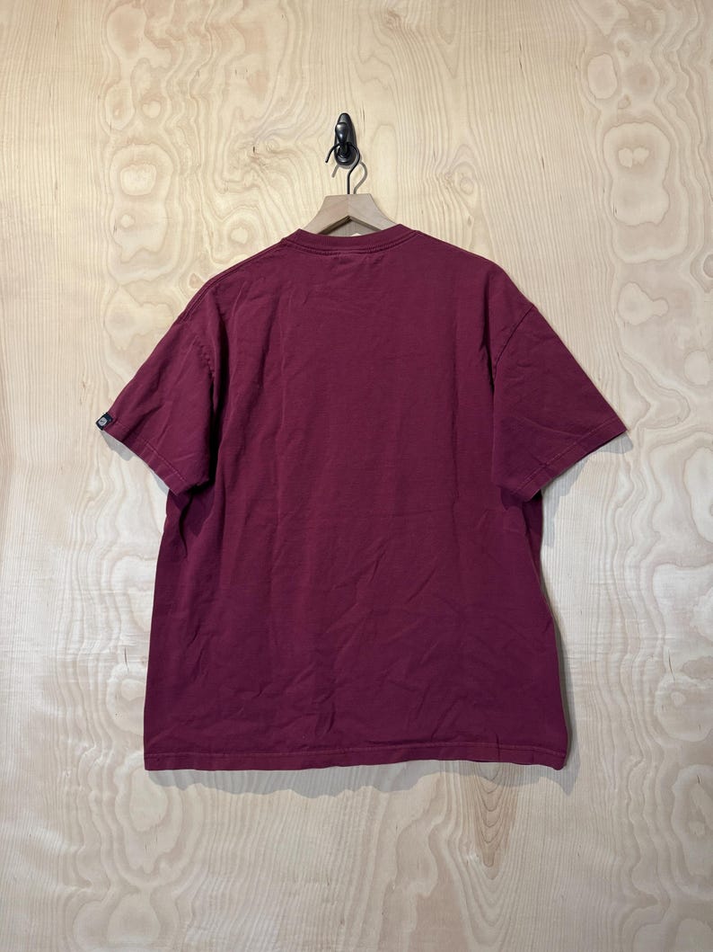 K&ouml;nnte beinhalten: Ein weinrotes T-Shirt mit kurzen &Auml;rmeln h&auml;ngt an einem Holzkleiderb&uuml;gel. Das Shirt besteht aus einem weichen Material und hat ein schlichtes, klassisches Design. Der Hintergrund ist eine hellfarbige Holzoberfl&auml;che.