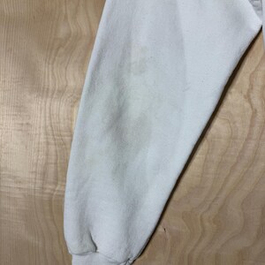 May include: Close-up of a cream-colored sweatshirt sleeve. The sleeve is slightly wrinkled and shows some discoloration. The cuff is ribbed and the fabric appears to be a soft, possibly fleece material. The background is a light wood grain.