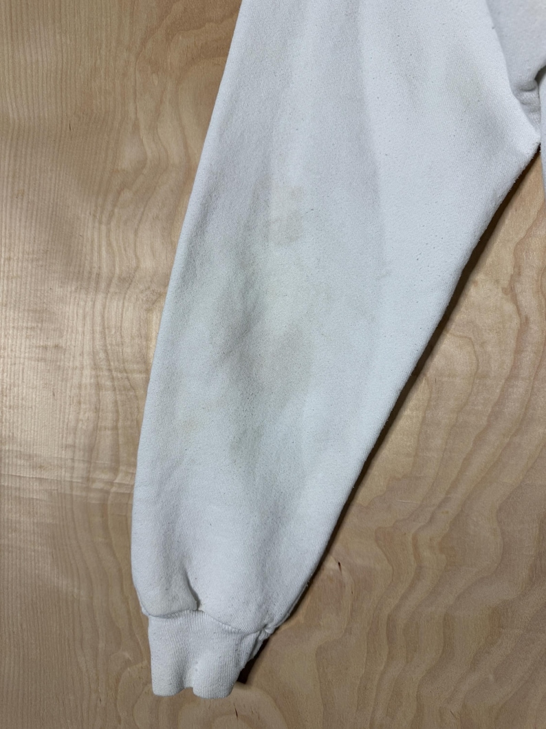 May include: Close-up of a cream-colored sweatshirt sleeve. The sleeve is slightly wrinkled and shows some discoloration. The cuff is ribbed and the fabric appears to be a soft, possibly fleece material. The background is a light wood grain.