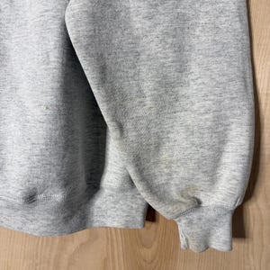K&ouml;nnte beinhalten: Nahaufnahme eines melierten grauen Sweatshirts. Das Bild zeigt den &Auml;rmel und einen Teil des K&ouml;rpers des Kleidungsst&uuml;cks. Die B&uuml;ndchen und der Saum sind gerippt. Der Stoff hat ein gesprenkeltes Aussehen. Es gibt einige Flecken auf dem &Auml;rmel.