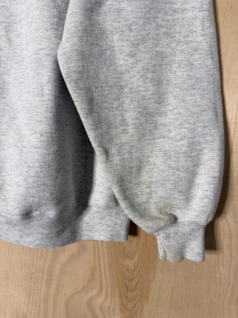 K&ouml;nnte beinhalten: Nahaufnahme eines melierten grauen Sweatshirts. Das Bild zeigt den &Auml;rmel und einen Teil des K&ouml;rpers des Kleidungsst&uuml;cks. Die B&uuml;ndchen und der Saum sind gerippt. Der Stoff hat ein gesprenkeltes Aussehen. Es gibt einige Flecken auf dem &Auml;rmel.