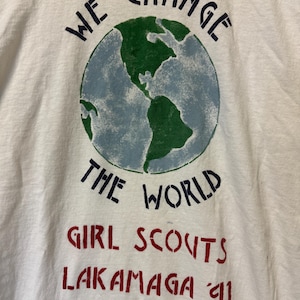 Peut inclure: T-shirt blanc avec un motif de globe terrestre vert et bleu. Le texte "WE CHANGE THE WORLD" est au-dessus du globe. En dessous, le texte "GIRL SCOUTS LAKAMAGA '91" est en rouge.