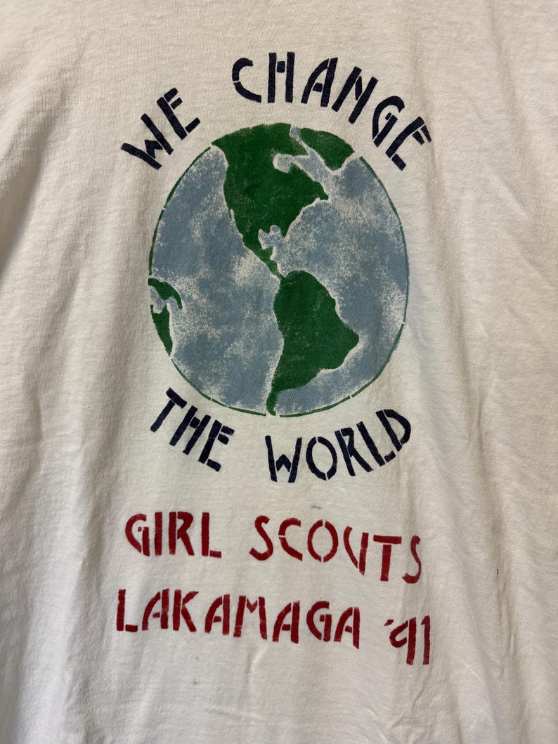 Peut inclure: T-shirt blanc avec un motif de globe terrestre vert et bleu. Le texte "WE CHANGE THE WORLD" est au-dessus du globe. En dessous, le texte "GIRL SCOUTS LAKAMAGA '91" est en rouge.