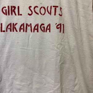 Peut inclure: T-shirt blanc avec des lettres bleues et rouges. Le texte indique "THE WORLD GIRL SCOUTS LAKAMAGA '91". Le t-shirt semble vintage et pr&eacute;sente une l&eacute;g&egrave;re d&eacute;coloration.