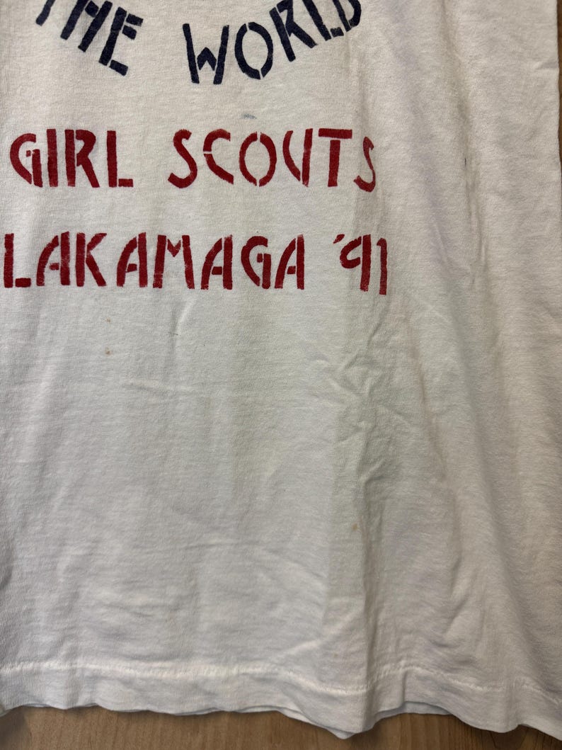 Peut inclure: T-shirt blanc avec des lettres bleues et rouges. Le texte indique "THE WORLD GIRL SCOUTS LAKAMAGA '91". Le t-shirt semble vintage et pr&eacute;sente une l&eacute;g&egrave;re d&eacute;coloration.