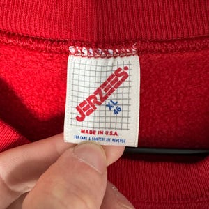 May include: A close-up of a red sweatshirt with a white label. The label displays "JERZEES" in red, with "XL 46" and "MADE IN U.S.A." printed below. The sweatshirt is suspended on a wooden hanger.