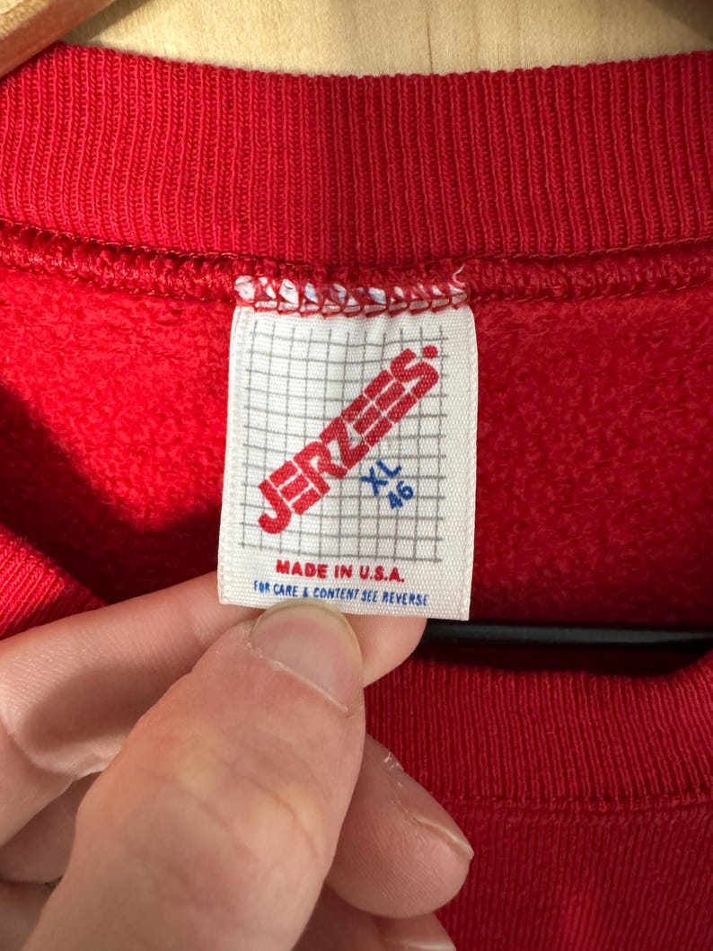 May include: A close-up of a red sweatshirt with a white label. The label displays "JERZEES" in red, with "XL 46" and "MADE IN U.S.A." printed below. The sweatshirt is suspended on a wooden hanger.
