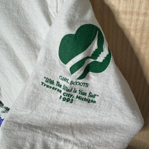 May include: White t-shirt sleeve featuring a green Girl Scouts logo and text. The text reads "GIRL SCOUTS", "With The Wind In Your Sail", "Traverse City, Michigan 1993". The logo is a green trefoil with a profile silhouette.