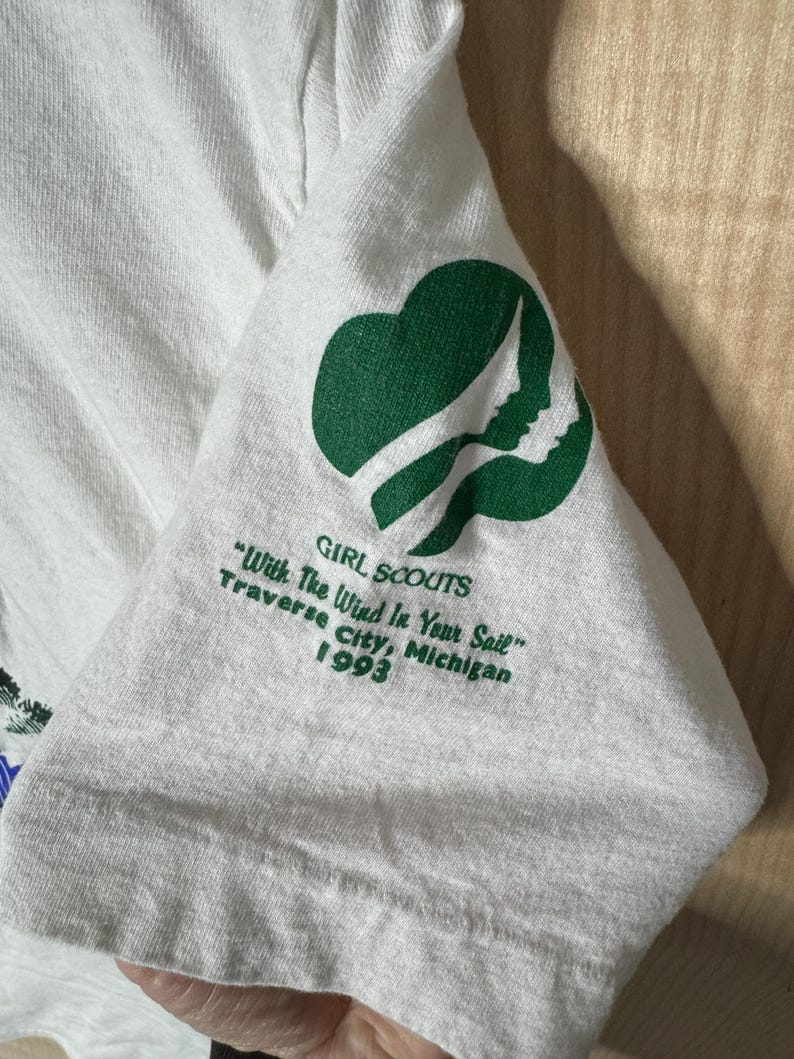 May include: White t-shirt sleeve featuring a green Girl Scouts logo and text. The text reads "GIRL SCOUTS", "With The Wind In Your Sail", "Traverse City, Michigan 1993". The logo is a green trefoil with a profile silhouette.