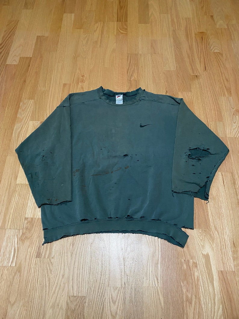 May include: A green sweatshirt with a distressed look, featuring a small Nike logo on the left chest. The sweatshirt has several holes and frayed edges.