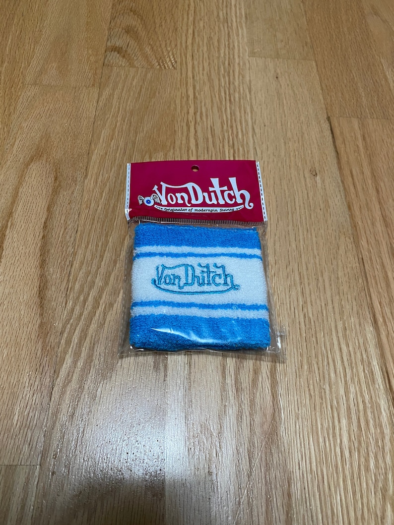 Peut inclure: Un bracelet de poignet ray&eacute; bleu et blanc avec le logo Von Dutch brod&eacute; en turquoise. Le bracelet est emball&eacute; dans un sac en plastique transparent avec une carte d'en-t&ecirc;te rouge et blanche qui indique "Von Dutch Originator of modernpin stripes".