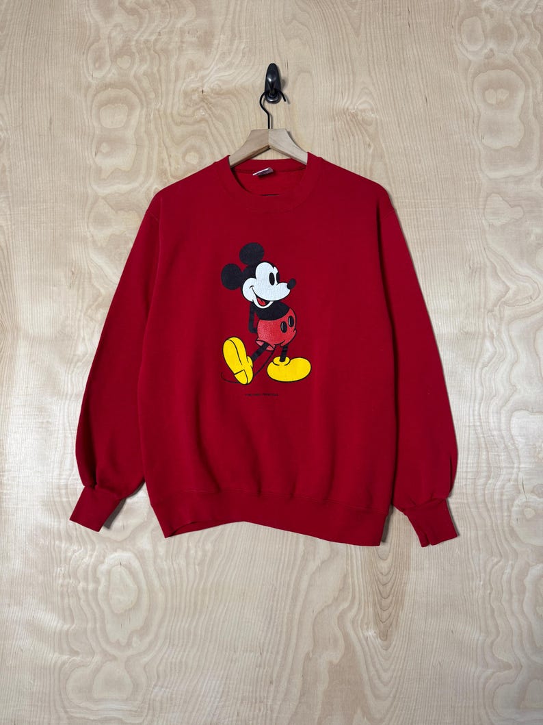 May include: Red sweatshirt with a Mickey Mouse graphic. The cartoon character is shown in his iconic outfit: black ears, white face, red shorts, yellow shoes, and white gloves. The long-sleeved top is suspended from a wooden hanger.