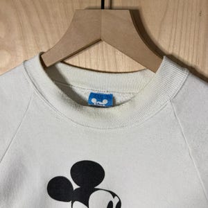 May include: Cream-colored sweatshirt featuring a large graphic of Mickey Mouse. The black and white cartoon character has a red and yellow outfit. The sweatshirt has a crew neck and long sleeves. A blue tag with the Disney logo is visible.