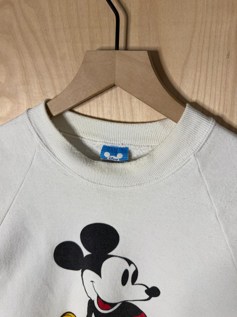 May include: Cream-colored sweatshirt featuring a large graphic of Mickey Mouse. The black and white cartoon character has a red and yellow outfit. The sweatshirt has a crew neck and long sleeves. A blue tag with the Disney logo is visible.