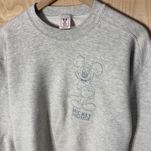 K&ouml;nnte beinhalten: Ein hellgraues Mickey Mouse Sweatshirt mit Rundhalsausschnitt. Das Sweatshirt hat ein blaues, gesticktes Bild von Mickey Mouse auf der Vorderseite mit den Worten "Mickey Mouse" darunter. Das Disney-Logo ist auf dem Etikett sichtbar.