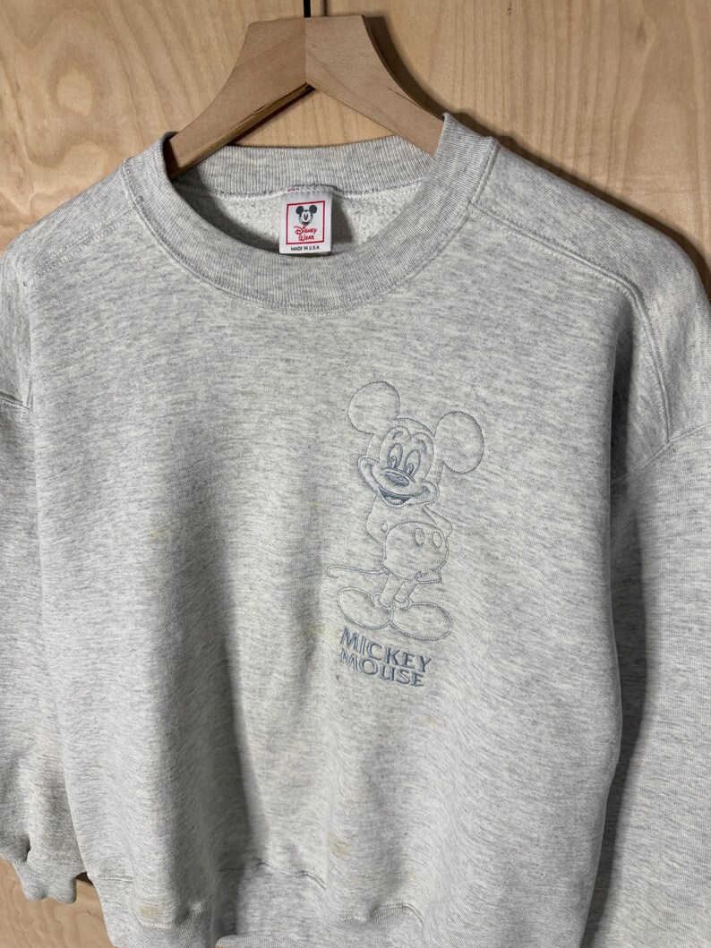 K&ouml;nnte beinhalten: Ein hellgraues Mickey Mouse Sweatshirt mit Rundhalsausschnitt. Das Sweatshirt hat ein blaues, gesticktes Bild von Mickey Mouse auf der Vorderseite mit den Worten "Mickey Mouse" darunter. Das Disney-Logo ist auf dem Etikett sichtbar.