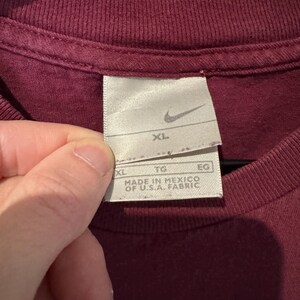K&ouml;nnte beinhalten: Nahaufnahme eines weinroten T-Shirts mit einem Nike-Etikett. Das Etikett ist wei&szlig; mit dem Nike-Swoosh-Logo und der Gr&ouml;&szlig;e "XL". Das Etikett lautet ebenfalls "MADE IN MEXICO OF U.S.A. FABRIC".