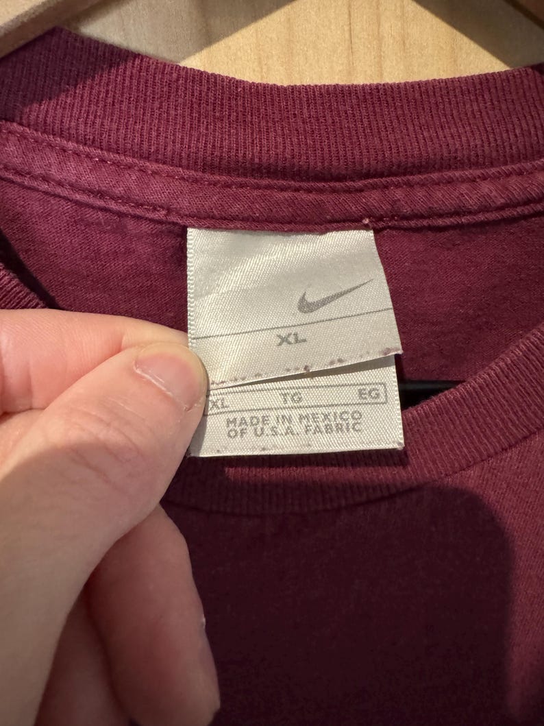 K&ouml;nnte beinhalten: Nahaufnahme eines weinroten T-Shirts mit einem Nike-Etikett. Das Etikett ist wei&szlig; mit dem Nike-Swoosh-Logo und der Gr&ouml;&szlig;e "XL". Das Etikett lautet ebenfalls "MADE IN MEXICO OF U.S.A. FABRIC".