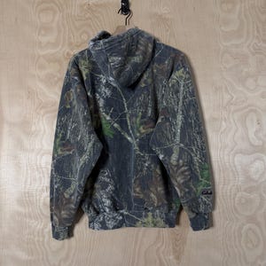 May include: A long-sleeved hooded sweatshirt featuring a dark forest camouflage pattern with shades of grey, brown, and green. The garment includes a hood, ribbed cuffs, a ribbed hem, and the Cabela's brand name embroidered on the left sleeve.