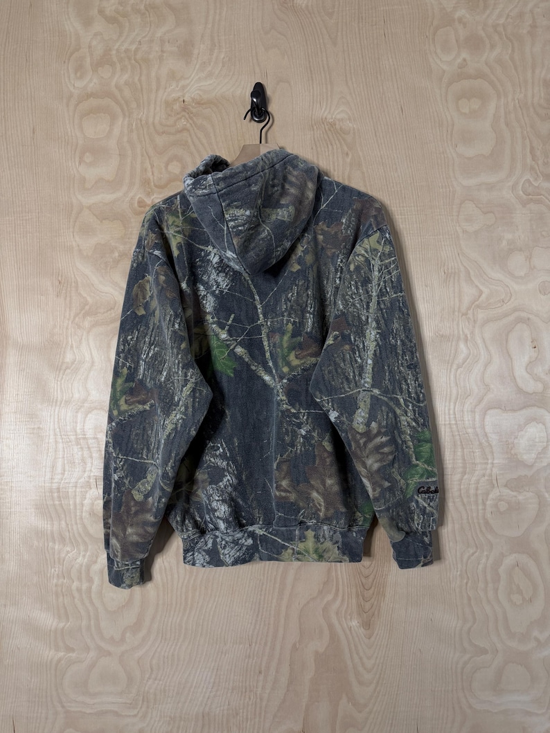 May include: A long-sleeved hooded sweatshirt featuring a dark forest camouflage pattern with shades of grey, brown, and green. The garment includes a hood, ribbed cuffs, a ribbed hem, and the Cabela's brand name embroidered on the left sleeve.