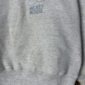 K&ouml;nnte beinhalten: Hellgraues Sweatshirt mit gesticktem Mickey Mouse-Motiv. Der Schriftzug "MICKEY MOUSE" ist darunter in Blau gestickt. Das Sweatshirt hat einen Rundhalsausschnitt und gerippte B&uuml;ndchen, typisch f&uuml;r Freizeitkleidung.