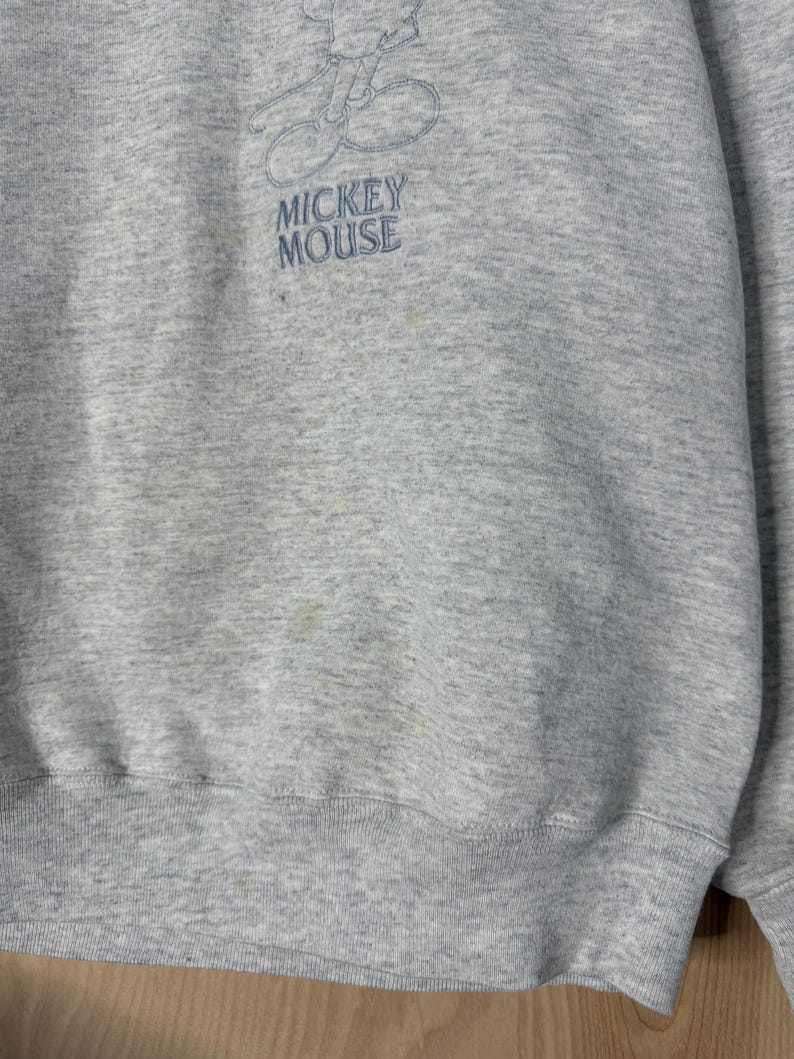 K&ouml;nnte beinhalten: Hellgraues Sweatshirt mit gesticktem Mickey Mouse-Motiv. Der Schriftzug "MICKEY MOUSE" ist darunter in Blau gestickt. Das Sweatshirt hat einen Rundhalsausschnitt und gerippte B&uuml;ndchen, typisch f&uuml;r Freizeitkleidung.