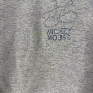 K&ouml;nnte beinhalten: Ein graues Sweatshirt mit einem verblassten, gestickten Bild von Mickey Mouse. Der Text "MICKEY MOUSE" ist unter der Figur gestickt. Der Stoff weist Gebrauchsspuren und Verf&auml;rbungen auf.