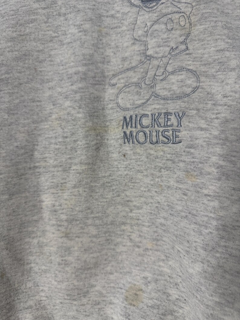 K&ouml;nnte beinhalten: Ein graues Sweatshirt mit einem verblassten, gestickten Bild von Mickey Mouse. Der Text "MICKEY MOUSE" ist unter der Figur gestickt. Der Stoff weist Gebrauchsspuren und Verf&auml;rbungen auf.