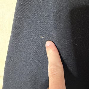May include: Close-up of a dark gray fabric with a textured surface. A finger is pointing at the fabric. The fabric appears to be a knit or woven material, possibly cotton or a blend.