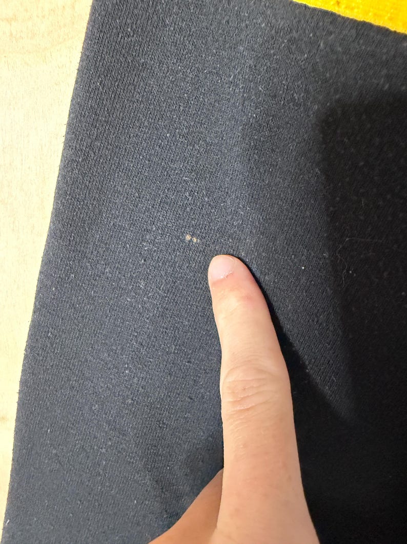 May include: Close-up of a dark gray fabric with a textured surface. A finger is pointing at the fabric. The fabric appears to be a knit or woven material, possibly cotton or a blend.