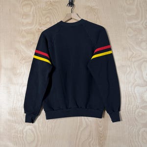 May include: Black sweatshirt with red and yellow stripes on the sleeves. The crew neck sweatshirt is hanging on a wooden hanger against a light wood grain background. The sweatshirt is a vintage style.