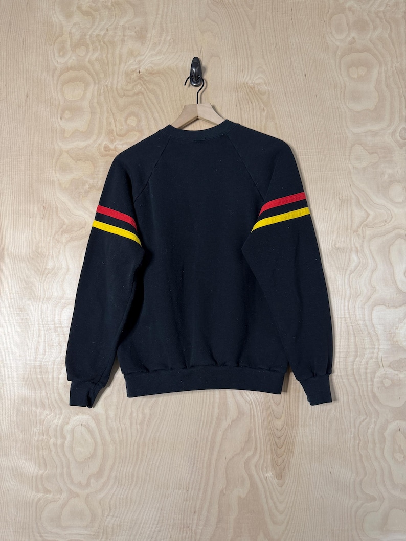 May include: Black sweatshirt with red and yellow stripes on the sleeves. The crew neck sweatshirt is hanging on a wooden hanger against a light wood grain background. The sweatshirt is a vintage style.