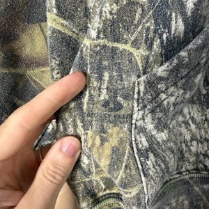 May include: A hooded sweatshirt featuring a camouflage pattern in shades of dark grey, olive green, and muted brown. The garment includes a front kangaroo pocket and ribbed cuffs, showcasing a detailed forest-inspired print across the fabric.