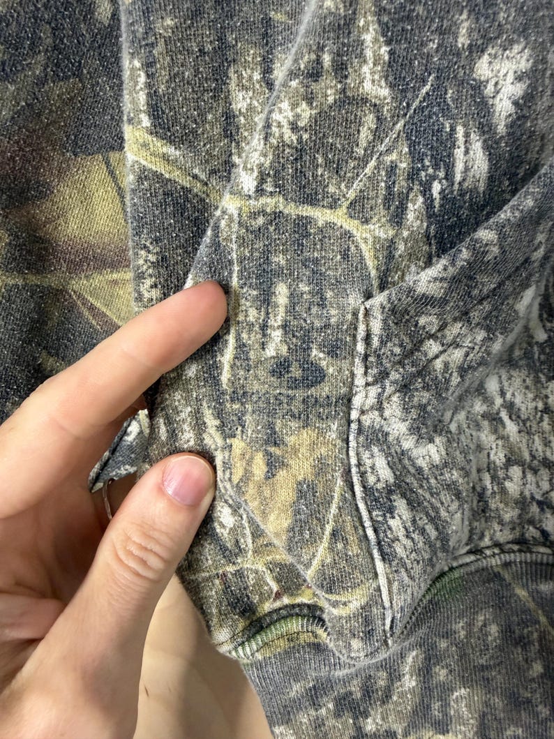 May include: A hooded sweatshirt featuring a camouflage pattern in shades of dark grey, olive green, and muted brown. The garment includes a front kangaroo pocket and ribbed cuffs, showcasing a detailed forest-inspired print across the fabric.