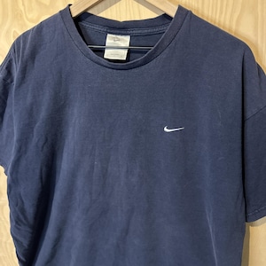 May include: Navy blue t-shirt featuring a white Nike swoosh logo. The shirt is displayed on a wooden hanger. The fabric has a faded appearance, possibly indicating a vintage or pre-worn garment.