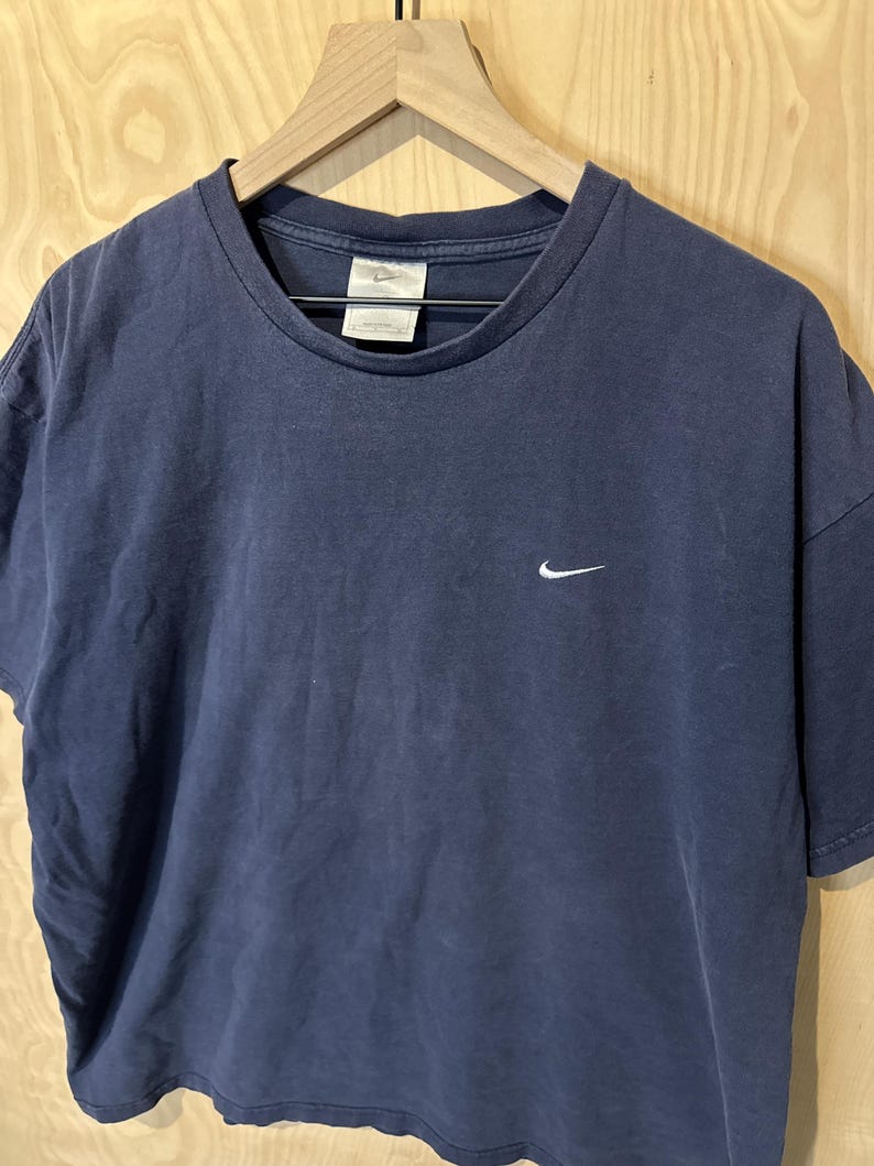 May include: Navy blue t-shirt featuring a white Nike swoosh logo. The shirt is displayed on a wooden hanger. The fabric has a faded appearance, possibly indicating a vintage or pre-worn garment.