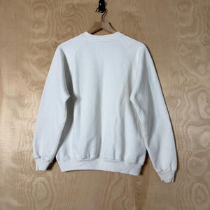 May include: A plain white sweatshirt with long sleeves, hanging on a wooden hanger. The sweatshirt is made of a soft material and has a crew neck. The background is a light wood grain.