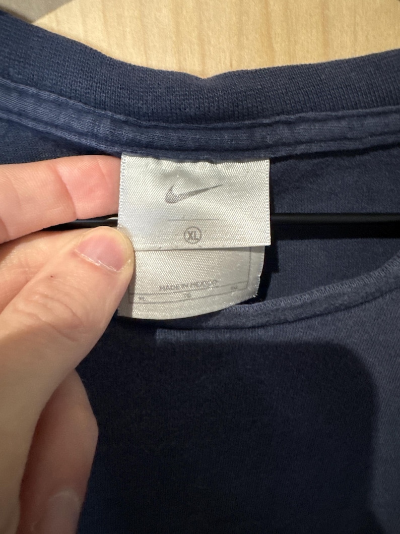 May include: Close-up of a navy blue t-shirt with a white tag. The tag features the Nike logo and the size XL. The tag also includes the text "MADE IN MEXICO".