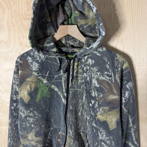 May include: A full-zip hooded sweatshirt featuring a forest camouflage pattern in shades of grey, brown, and green. The garment includes a drawstring hood, a central black zip closure, and two front pockets. The Cabela's brand label is visible.