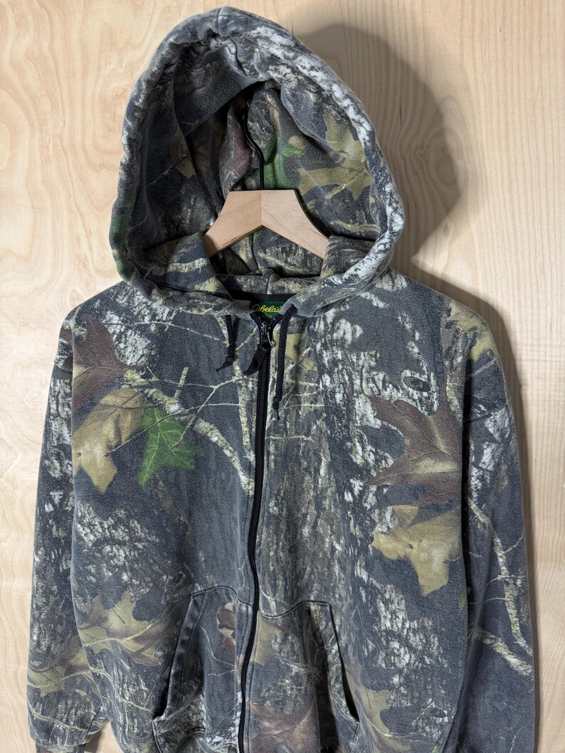 May include: A full-zip hooded sweatshirt featuring a forest camouflage pattern in shades of grey, brown, and green. The garment includes a drawstring hood, a central black zip closure, and two front pockets. The Cabela's brand label is visible.