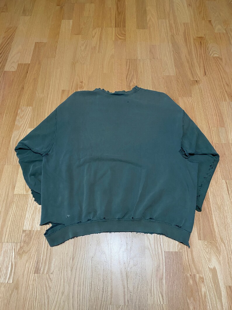 May include: A dark green sweatshirt with a distressed, worn look. The sweatshirt has a crew neck and long sleeves. The fabric has a soft, comfortable feel.