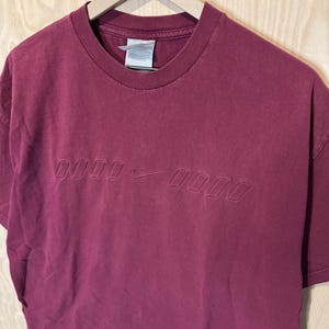 K&ouml;nnte beinhalten: Ein weinrotes T-Shirt mit Rundhalsausschnitt und kurzen &Auml;rmeln. Das Shirt hat ein gepr&auml;gtes Design mit dem Nike Swoosh-Logo und Text. Es h&auml;ngt an einem Holzkleiderb&uuml;gel.