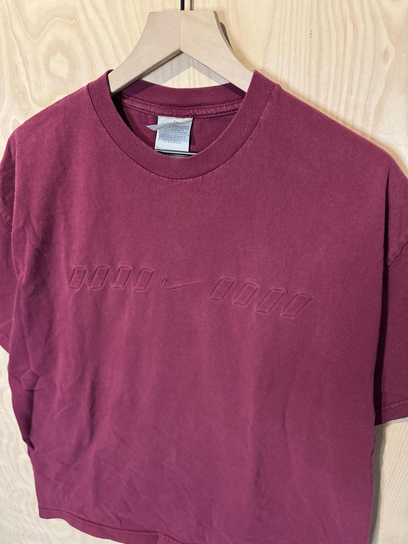 K&ouml;nnte beinhalten: Ein weinrotes T-Shirt mit Rundhalsausschnitt und kurzen &Auml;rmeln. Das Shirt hat ein gepr&auml;gtes Design mit dem Nike Swoosh-Logo und Text. Es h&auml;ngt an einem Holzkleiderb&uuml;gel.
